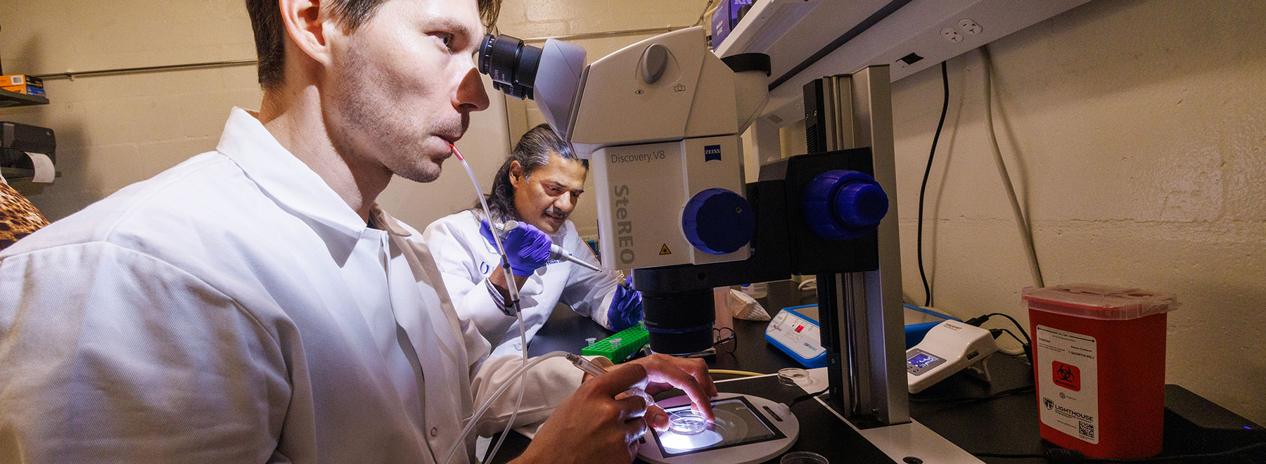 closeup of two researchers at work in the laboratory. The researcher in the foreground uses a device controlled by mouth